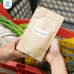Mains tenant un sachet kraft portant une étiquette Tea leaf, devant un cageot de légumes frais