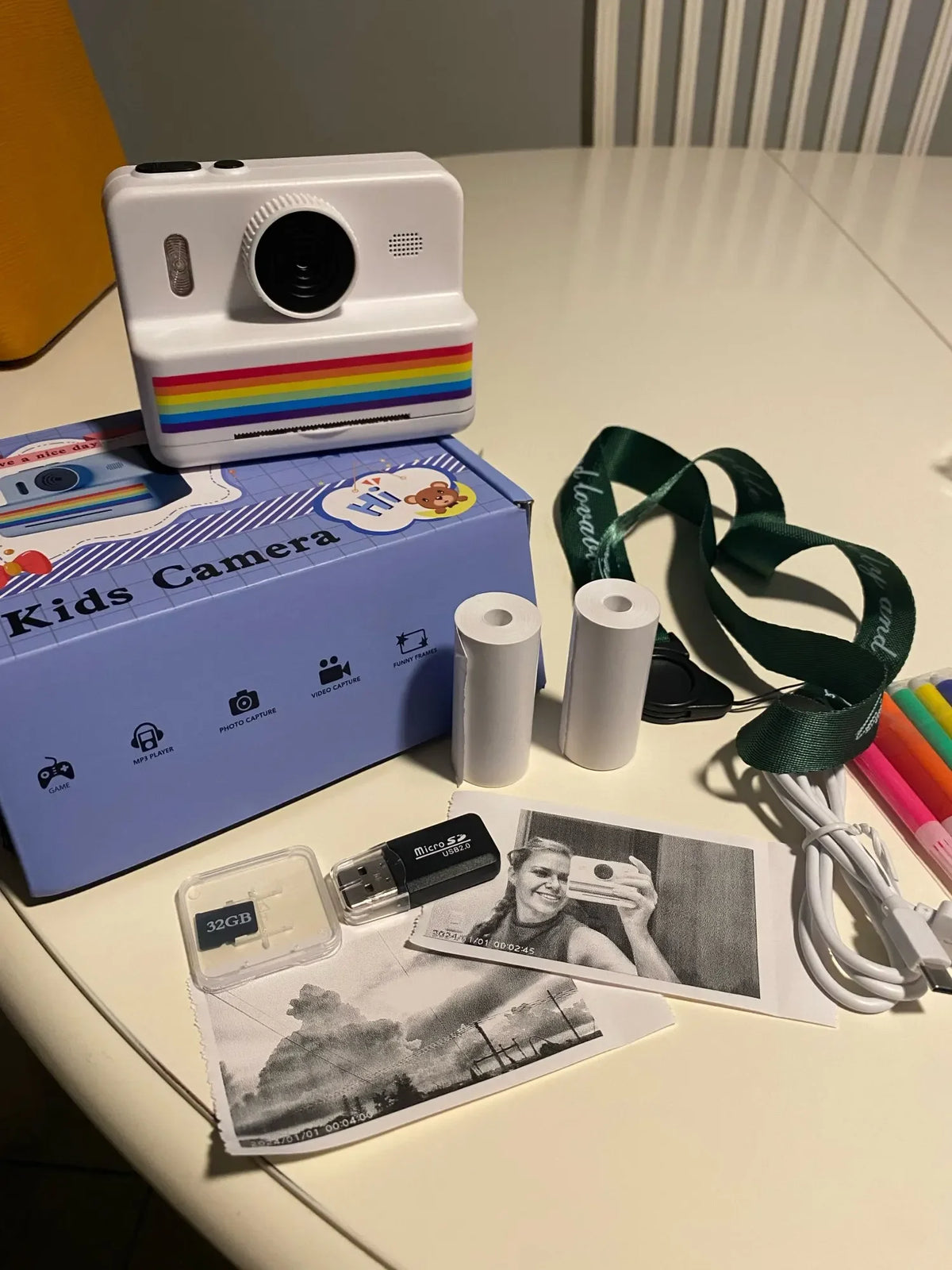 Déballage sur une table : boîte Kids Camera violette, rouleaux de papier, dragonne, carte 32 Go et câble