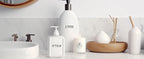 Salle de bains avec flacons étiquetés shampooing et gel douche datés, bougie et diffuseur sur le marbre